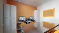 This business center is a well-appointed, single-level workspace designed to meet a range of professional needs. The room's most distinctive design element is a bold orange chevron-patterned accent wall, which provides a vibrant contrast against the surrounding white walls and crown molding. The flooring throughout is warm-toned hardwood, lending a polished yet inviting atmosphere to the space.

A built-in workstation along the accent wall accommodates two desktop computers and a multifunction printer, supported by white cabinetry that provides ample built-in storage above and below. Yellow upholstered chairs add a coordinated pop of color at both the workstation and the central glass-top table, which serves as an additional workspace or informal meeting area.

Cork bulletin boards with orange frames are mounted on the white walls, offering practical display and note-taking surfaces. A large window fitted with horizontal blinds allows natural light to filter into the room while maintaining privacy, with views of surrounding greenery outside. The space is accessed through a classic French-paned door with a matte black handle, complemented by wainscoting in the adjacent hallway, where framed artwork adds a finishing decorative touch.