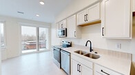 This thoughtfully designed studio unit at 220 Quimby St NE in Grand Rapids, Michigan offers a compact yet highly functional living space across a single floor. The open-concept layout seamlessly connects the kitchen and living area, maximizing every square foot. Light-toned laminate flooring runs throughout, lending a clean and cohesive look to the entire unit.

The kitchen is a standout feature, outfitted with crisp white shaker cabinetry with matte black hardware, light-colored countertops, and a full suite of stainless steel appliances including a refrigerator, electric range, dishwasher, and over-the-range microwave. A double-basin sink with a black faucet completes the well-appointed cooking space.

The main living area benefits from abundant natural light through a large sliding glass door that opens to a private balcony, offering outdoor views of the surrounding neighborhood. A striking matte black sliding barn door adds a modern industrial accent and provides flexible room division within the space.

The bathroom is clean and functional, featuring a white vanity with matching hardware, recessed lighting, and a walk-in shower area. Additional coat hooks and storage nooks are built into the bathroom corridor. Recessed ceiling lighting throughout keeps the interior bright and contemporary. This unit presents a polished, move-in-ready living experience in a modern residential building.