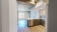Located at 800 S Western Ave in Los Angeles, this modern one-bedroom, one-bathroom unit blends industrial-chic aesthetics with contemporary finishes throughout. The open-concept kitchen and living area serves as the heart of the space, showcasing striking exposed concrete beams and columns that lend an urban loft character. The kitchen is well-appointed with two-tone cabinetry — light wood uppers paired with charcoal lowers — white quartz countertops, a tile backsplash, stainless steel appliances including a refrigerator, range, microwave, and dishwasher, and pendant lighting that adds warmth overhead. Light wood-tone flooring flows seamlessly throughout the unit. The living area benefits from natural light through windows offering city views. The bedroom is a bright, tranquil retreat featuring exposed concrete ceiling details, a large window with urban views, and a sliding double-door closet. The hallway connects all spaces efficiently, with a louvered interior door adding a design accent. The bathroom is clean and modern, featuring light stone-look tile flooring, a floating gray vanity, white countertop, and an illuminated frameless mirror. This well-designed unit delivers a sophisticated urban living experience in the heart of Los Angeles.