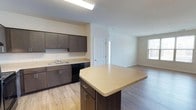 This single-floor space offers a clean, modern aesthetic defined by an open-concept layout that seamlessly connects the kitchen and living area. The kitchen is well-appointed with dark espresso-toned cabinetry providing generous storage, complemented by light quartz-style countertops that offer ample workspace. A central island extends the counter space further, making it ideal for meal preparation or casual dining. Black appliances, including a dishwasher, add a sleek, contemporary contrast to the cabinetry. A stainless steel sink sits beneath overhead task lighting, ensuring a bright and functional workspace. The flooring throughout is a warm, light-toned hardwood-style material that adds continuity and warmth across the entire space. The living area flows naturally from the kitchen, bathed in natural light from a window that brightens the room and creates an airy, welcoming atmosphere. Neutral white walls throughout keep the space feeling open and versatile, ready to accommodate a variety of interior styles. The overall ambiance is fresh and move-in ready, reflecting thoughtful design choices that balance practicality with modern appeal.