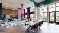 Located at 7222 S Peek Rd in Houston, TX, this single-floor community clubhouse delivers a striking contemporary design defined by soaring double-height ceilings and an energetic, color-forward aesthetic. The expansive open floor plan is unified by a vivid geometric patchwork carpet that anchors the social zones, transitioning to a rich herringbone-patterned tile in the dining area. Bold architectural accents — including teal-painted upper walls, deep magenta columns, and textured feature walls with intricate geometric patterning — create a dynamic visual identity throughout the space. Floor-to-ceiling steel-framed windows flood the interior with abundant natural light while framing views of the surrounding community. A large communal dining table surrounded by plush velvet chairs provides a refined gathering spot, while the game area features a full-size billiards table illuminated by a statement ring pendant light. A dedicated lounge zone offers curved teal velvet seating arranged around a sculptural gold coffee table, ideal for casual conversation. Additional amenities include a foosball table and a media nook with a mounted display. The layered design elements, generous proportions, and curated furnishings combine to create an inviting, socially dynamic environment suited for both relaxed gatherings and lively entertainment.