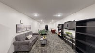 Located at 731 E Terra Cotta Ave in Crystal Lake, Illinois, this single-floor community living room is a thoughtfully designed multipurpose gathering space. The room is finished with crisp white walls and durable patterned carpet tiles that add texture and visual interest underfoot, while recessed ceiling lighting provides bright, even illumination throughout.

The space is organized into several distinct activity zones. A comfortable lounge area features a gray upholstered sofa paired with a sleek coffee table, ideal for relaxed conversation or quiet downtime. Adjacent to the lounge, a round dining table surrounded by green upholstered chairs offers a welcoming spot for group gatherings or casual games. A full-size shuffleboard table serves as a central recreational feature, adding an engaging social element to the room.

Along one wall, a media console supports a large flat-screen television, flanked by dark open bookshelves stocked with board games, books, and decorative greenery. Additional bookshelves line another section of the room, filled with a wide selection of reading material and accented with framed artwork on the walls. A cozy seating nook with barrel chairs and a small side table provides a quieter corner for reading or chess. The overall ambiance is clean, functional, and inviting, making this space well-suited for community relaxation and recreation.