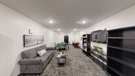 Located at 731 E Terra Cotta Ave in Crystal Lake, Illinois, this single-floor community living room is a thoughtfully designed multipurpose gathering space. The room is finished with crisp white walls and durable patterned carpet tiles that add texture and visual interest underfoot, while recessed ceiling lighting provides bright, even illumination throughout.

The space is organized into several distinct activity zones. A comfortable lounge area features a gray upholstered sofa paired with a sleek coffee table, ideal for relaxed conversation or quiet downtime. Adjacent to the lounge, a round dining table surrounded by green upholstered chairs offers a welcoming spot for group gatherings or casual games. A full-size shuffleboard table serves as a central recreational feature, adding an engaging social element to the room.

Along one wall, a media console supports a large flat-screen television, flanked by dark open bookshelves stocked with board games, books, and decorative greenery. Additional bookshelves line another section of the room, filled with a wide selection of reading material and accented with framed artwork on the walls. A cozy seating nook with barrel chairs and a small side table provides a quieter corner for reading or chess. The overall ambiance is clean, functional, and inviting, making this space well-suited for community relaxation and recreation.