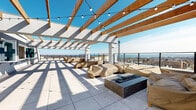 Located at 50 Lecount Place in New Rochelle, NY, this rooftop terrace offers an expansive open-air gathering space elevated above the city skyline. The terrace is defined by a striking wood-beam pergola structure with white paneled roofing sections, which provides partial shade while allowing abundant natural light to filter through. String lights are strung across the pergola beams, lending the space an inviting ambiance suited for both daytime and evening gatherings.

The large-format tile flooring extends throughout the terrace, creating a clean, modern aesthetic. A centrally positioned fire pit serves as a focal point for the main lounge area, which is furnished with multiple covered seating arrangements, including sofas and lounge chairs arranged to encourage social interaction. An additional seating alcove is situated near the pergola's interior wall, providing a more intimate setting.

The terrace is enclosed by sleek metal railings that preserve unobstructed panoramic views of the New Rochelle cityscape and the Long Island Sound beyond. Restroom facilities and utility access are available on-site, and a posted maximum occupancy of 150 persons underscores the terrace's capacity as a significant event and amenity space. A grilling station is also visible, further enhancing the terrace's functionality for outdoor entertaining.