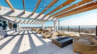 Located at 50 Lecount Place in New Rochelle, NY, this rooftop terrace offers an expansive open-air gathering space elevated above the city skyline. The terrace is defined by a striking wood-beam pergola structure with white paneled roofing sections, which provides partial shade while allowing abundant natural light to filter through. String lights are strung across the pergola beams, lending the space an inviting ambiance suited for both daytime and evening gatherings.

The large-format tile flooring extends throughout the terrace, creating a clean, modern aesthetic. A centrally positioned fire pit serves as a focal point for the main lounge area, which is furnished with multiple covered seating arrangements, including sofas and lounge chairs arranged to encourage social interaction. An additional seating alcove is situated near the pergola's interior wall, providing a more intimate setting.

The terrace is enclosed by sleek metal railings that preserve unobstructed panoramic views of the New Rochelle cityscape and the Long Island Sound beyond. Restroom facilities and utility access are available on-site, and a posted maximum occupancy of 150 persons underscores the terrace's capacity as a significant event and amenity space. A grilling station is also visible, further enhancing the terrace's functionality for outdoor entertaining.