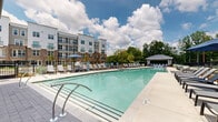 This expansive outdoor amenity space centers around a large, rectangular swimming pool with clear aquamarine water, designed to serve the residents of a modern multi-story building complex that forms an impressive backdrop. The pool deck is finished in light-toned concrete, providing ample space for relaxation and recreation. Rows of cushioned chaise lounges line both sides of the pool, offering plenty of room for sunbathing, and are complemented by shaded seating areas furnished with contemporary outdoor chairs and planters. A dedicated pool house structure sits at one end of the deck, featuring a covered porch and additional seating, and likely houses restrooms and storage facilities. At the opposite end, a smaller covered structure provides additional shade and convenience. The surrounding multi-story residential building showcases a modern architectural design, blending white paneling with warm brick accents, large windows, and private balconies that overlook the pool area. A black iron perimeter fence encloses the space for privacy and security, while mature trees and greenery beyond the fence contribute a natural, park-like ambiance. The entire outdoor area is bright and open, benefiting from abundant natural light and open sky views, creating an inviting resort-like atmosphere ideal for leisure and outdoor living.