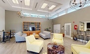 Located at 25001 SW 130th Ave in Princeton, FL, this single-floor community space offers a thoughtfully designed open-concept layout that seamlessly blends a living room and kitchen into one cohesive environment. The interior immediately impresses with its dramatically high ceilings, which are accentuated by a series of coffered panels and large skylights that flood the space with abundant natural light. Striking orb-style chandeliers hang at varying heights, adding an elegant yet contemporary focal point throughout the room. The living area features light wood-style tile flooring that runs throughout, creating a clean and unified aesthetic. Comfortable seating — including soft yellow accent chairs and neutral gray sofas — is arranged around a built-in wood media and shelving unit, providing both function and warmth. A dining area with dark wood tables and woven rattan chairs sits adjacent, separated by a textured wall panel that adds visual depth. Large glass doors and clerestory windows along the perimeter invite views of the surrounding outdoor areas and allow natural light to pour in from multiple angles. The entrance foyer area includes louvered doors and utility access points, maintaining a tidy and organized flow. Overall, the space exudes a bright, modern, and welcoming ambiance suited for community gathering and everyday use.