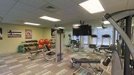 This fitness center is designed to support a comprehensive workout routine in a clean, functional environment. The space is finished with a drop ceiling fitted with recessed panel lighting that ensures bright, even illumination throughout. Patterned carpet tiles cover the floor, adding texture and a subtle design element to the room.

The facility is outfitted with a diverse selection of equipment to accommodate various training needs. A dedicated free weights area lines one wall, featuring a full rack of dumbbells alongside stability balls for core and flexibility work. Multiple weight benches and adjustable training stations are positioned throughout the floor, providing ample options for strength training.

On the cardio side, treadmills and upright exercise bikes are arranged to face large wall-mounted flat-screen televisions, allowing users to stay entertained during their workouts. Full-length mirrors span the walls, visually expanding the space and allowing exercisers to monitor their form.

Natural light filters in through windows on one side of the room, complementing the overhead lighting and contributing to the bright, energizing atmosphere. The overall layout is thoughtfully organized to maximize usable floor space while keeping equipment accessible and the environment open and inviting for users of all fitness levels.