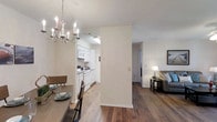 This single-floor residence offers a cohesive open layout that connects the dining, kitchen, and living areas in a practical and visually unified arrangement. The dining area is anchored by a rustic wood table and accented by a white candelabra-style chandelier that adds a touch of classic charm to the space. Hardwood-style flooring in warm tones flows consistently throughout, lending a sense of continuity and warmth across all visible areas.

The kitchen, partially separated by a structural wall, features white cabinetry, white appliances including a range and microwave, and light countertops that contribute to a clean, bright aesthetic. Natural and artificial lighting keeps the kitchen well-illuminated. A hallway leads toward the living area, which is softly lit by a table lamp and features a neutral-toned sofa, creating a comfortable and relaxed atmosphere. The walls throughout are painted in a crisp, neutral tone, enhancing the brightness of the space. The textured ceiling adds a subtle traditional detail. Overall, the residence presents a functional and inviting layout with consistent finishes throughout.