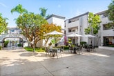 Located at 12200 Valley View St in Garden Grove, California, this Mediterranean-inspired property presents a cohesive architectural design characterized by white stucco exteriors, decorative arched entryways, wrought iron balcony railings, and clay tile roof accents. The multi-building complex is centered around a generously sized open courtyard, finished with light-toned stone pavers that extend throughout the outdoor common areas.

The courtyard serves as the social heart of the property, furnished with multiple umbrella-shaded dining tables and chairs, offering comfortable spaces for outdoor gatherings. Mature trees, including palms and broadleaf varieties, provide natural shade and visual interest, while manicured ground-level plantings and garden beds add vibrancy throughout. Classic black lamppost fixtures line the walkways, contributing to the property's charming aesthetic.

Arched passageways connect various sections of the complex, enhancing the Spanish Colonial character of the design. Individual unit entrances feature private covered patio areas with ground-level garden borders, offering a sense of personal outdoor space. Large sliding glass doors allow for natural light and views of the surrounding greenery. The well-maintained landscaping and cohesive architectural details create an inviting, resort-like ambiance throughout this Southern California property.