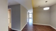 This single-floor space showcases a clean, modern design characterized by rich, dark hardwood-style flooring that runs throughout the interior, creating a warm and cohesive aesthetic. The walls are finished in neutral tones — a combination of cool gray and warm taupe — providing a versatile backdrop that complements a variety of décor styles. White baseboards offer a crisp contrast against the darker flooring, adding a polished, finished look to the space.

A central hallway connects the entry area to the main living space, which is visible at the far end and benefits from abundant natural light streaming through large windows. The bright, open atmosphere of the far room stands in pleasing contrast to the more enclosed entryway corridor, creating a natural sense of flow and transition through the space.

Architectural details include a ceiling-mounted HVAC vent, indicating a centralized climate control system, as well as standard electrical outlets along the walls. A louvered door, likely for a closet, is visible along the hallway, adding practical storage to the layout. Overall, the space presents a well-maintained, move-in-ready environment with a modern sensibility.