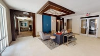 This contemporary clubroom is designed to offer residents a sophisticated and versatile social space across a single, well-appointed floor. The main lounge area immediately draws attention with its striking suspended ceiling feature — a dark wood-framed panel adorned with an intricate geometric lattice design — anchored by a modern chandelier. A bold teal accent wall adds a vibrant pop of color, complementing the neutral large-format tile flooring. Comfortable seating arrangements, including a plush sofa and accent chairs, are centered on an area rug, creating an inviting gathering space. Glass double doors open to an outdoor patio, allowing natural light to fill the room.

Adjacent to the lounge, a full bar area features a wide white quartz countertop with a vivid teal island base, wood-plank accent tiling along the back wall, and pendant lighting overhead — ideal for entertaining. Rustic wood bar stools complete the casual yet refined aesthetic.

The game room transitions seamlessly from the lounge via warm hardwood flooring and offers a full-sized billiards table illuminated by a sleek industrial-style pendant fixture, a built-in tufted banquette for spectators, a shuffleboard table, and a comfortable seating nook. Abstract artwork and sculptural wall décor throughout the space add artistic character. Cove lighting and recessed fixtures provide layered illumination across every zone.