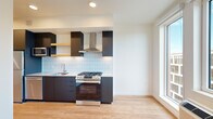 This thoughtfully designed single-floor unit offers a compact yet functional layout that makes efficient use of every square foot. The open living and kitchen area is anchored by a striking modern kitchen featuring dark cabinetry, light blue subway tile backsplash, stainless steel appliances including a gas range with a stainless hood, dishwasher, and refrigerator. A stacked in-unit washer and dryer is neatly integrated alongside the kitchen cabinetry, adding everyday convenience. A large floor-to-ceiling window floods the living area with abundant natural light and provides views of the surrounding neighborhood, enhancing the sense of openness throughout. Light hardwood flooring runs seamlessly across the entire unit, creating a warm and cohesive aesthetic. The bedroom area is served by a ceiling fan and features a spacious closet with open shelving and hanging rods, offering generous storage. The full bathroom is clean and bright, finished in white with a soaking tub and shower combo, a sleek gray vanity with an integrated sink, and recessed lighting. A connecting hallway with additional built-in shelving ties the layout together efficiently. The overall design balances modern finishes with a functional, minimalist sensibility.