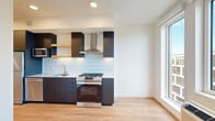 This thoughtfully designed single-floor unit offers a compact yet functional layout that makes efficient use of every square foot. The open living and kitchen area is anchored by a striking modern kitchen featuring dark cabinetry, light blue subway tile backsplash, stainless steel appliances including a gas range with a stainless hood, dishwasher, and refrigerator. A stacked in-unit washer and dryer is neatly integrated alongside the kitchen cabinetry, adding everyday convenience. A large floor-to-ceiling window floods the living area with abundant natural light and provides views of the surrounding neighborhood, enhancing the sense of openness throughout. Light hardwood flooring runs seamlessly across the entire unit, creating a warm and cohesive aesthetic. The bedroom area is served by a ceiling fan and features a spacious closet with open shelving and hanging rods, offering generous storage. The full bathroom is clean and bright, finished in white with a soaking tub and shower combo, a sleek gray vanity with an integrated sink, and recessed lighting. A connecting hallway with additional built-in shelving ties the layout together efficiently. The overall design balances modern finishes with a functional, minimalist sensibility.