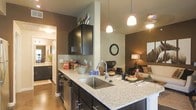 This single-floor residence showcases a thoughtfully designed open-concept layout that integrates the kitchen, dining, and living areas into one cohesive and inviting space. The kitchen serves as a focal point, featuring dark espresso cabinetry with sleek hardware, a granite countertop island with an undermount stainless steel sink, and a mosaic tile backsplash that adds a refined textural detail. Pendant lighting hangs above the island, casting a warm, ambient glow that complements the overall color palette of rich earth tones and neutral walls. The kitchen flows naturally into the living area, which is bathed in warm lighting and accented by decorative touches that enhance its cozy yet sophisticated ambiance. A hallway visible from the kitchen hints at additional rooms beyond, including what appears to be a well-appointed bathroom with light cabinetry and ample counter space. The polished concrete-style flooring runs throughout, providing a durable and modern foundation that ties the entire space together. The combination of dark cabinetry, granite surfaces, warm lighting, and an open floor plan makes this residence both functional and visually appealing.