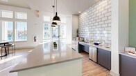 Located at 225 Rhyne Rd in Charlotte, NC, this single-floor community clubhouse offers a thoughtfully designed gathering space that blends modern aesthetics with everyday functionality. The open-concept layout is anchored by soaring ceilings and an abundance of natural light streaming through large clerestory and floor-to-ceiling windows, many of which frame views of the outdoor pool area.

The main lounge area features warm wood-look flooring, a stylish console table, and comfortable seating arrangements accented by bold orange armchairs — creating an inviting, social atmosphere. A full-length island kitchenette serves as a central hub, finished with sleek countertops, pendant lighting, stainless steel appliances, and a striking white subway tile backsplash wall that adds texture and depth to the space.

Adjacent to the kitchenette, a dedicated workspace area offers a quieter environment with carpeted flooring, built-in desk stations, and ample natural light from exterior-facing windows. The entryway and corridors are finished in a soft sage green, complemented by framed artwork and glass-paneled doors that maintain an open, airy feel throughout. Recessed lighting and clean white walls complete the polished, contemporary look, making this a versatile and welcoming community destination.