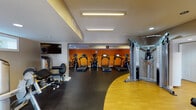 This fitness center is thoughtfully designed to accommodate a wide range of workout needs within a single-level layout. The space is divided into distinct functional zones, each dedicated to a specific type of training. The cardio area is lined with multiple treadmills and elliptical machines positioned against a bold orange accent wall, creating an energizing atmosphere. Overhead, linear fluorescent lighting ensures the area is well-illuminated throughout.

The strength-training zone features a selection of cable machines, selectorized weight stations, and isolated muscle-targeting equipment, all set on durable dark rubber flooring ideal for heavy use. A curved transition of warm hardwood-style flooring delineates this area, adding a visual contrast that helps define the space.

A separate section houses recumbent bikes and additional cardio equipment alongside a dumbbell rack, offering further versatility. Wall-mounted flat-screen televisions are positioned throughout for entertainment during workouts.

Near the entrance, stability balls are neatly stored on a vertical rack, and a lat pulldown station provides additional functional training options. Natural light filters in through small horizontal windows, complementing the bright overhead fixtures. The overall design balances practicality with an motivating aesthetic, making this fitness center a comprehensive facility for all fitness levels.