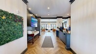 Located at 1407 S Grand Ave in Ames, Iowa, this expansive single-story amenity center spans over 5,000 square feet and offers a thoughtfully designed collection of spaces for socializing, fitness, and relaxation. The entrance greets visitors with herringbone-patterned hardwood flooring, bold dark trim, and recessed lighting that sets an upscale yet inviting tone throughout.

The centerpiece of the facility is a generously sized bar area featuring a long, dark-topped counter with leather bar stools, pendant lighting, and full service equipment. Adjacent lounge seating areas include bistro tables and chairs positioned beside floor-to-ceiling gridded windows overlooking an outdoor pool, allowing abundant natural light to fill the space.

A cozy lounge room with a painted brick feature wall, a wood-burning fireplace, dual wall-mounted TVs, and herringbone hardwood flooring offers a comfortable gathering spot. Dramatic barn-style sliding doors connect this area to a bright sitting lounge furnished with plush sofas and accented by tall windows and chandelier lighting.

The fitness facilities are divided across two rooms: one dedicated to free weights, benches, and functional training equipment, and a second outfitted with cable machines, a squat rack, and mirrored walls. Both rooms feature rubber flooring and dark-painted walls for a focused workout environment. A hallway connects all areas seamlessly throughout this well-appointed community amenity space.