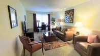 This single-floor space centers around a thoughtfully arranged living area that balances comfort and functionality. The main living space is anchored by a leather sofa and coordinating upholstered armchairs, arranged around a dark wood-and-glass coffee table atop a richly patterned area rug. Carpeted flooring in a neutral tone runs throughout, contributing to a warm and inviting ambiance.

Natural light filters in through a window dressed with deep burgundy curtains, brightening the room and adding a touch of color contrast against the light-colored walls. A decorative, multi-toned mosaic wall art piece serves as a focal point, adding visual interest and texture to the space.

The living area seamlessly transitions into a dedicated workspace, furnished with a dark desk and matching chairs, making the space adaptable for both relaxation and productivity. A tall indoor plant near the workspace introduces a natural element, softening the overall aesthetic. Ambient lighting from strategically placed floor and table lamps enhances the warm atmosphere throughout the room.