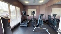 This single-level fitness room is designed to provide a functional and comfortable workout environment. The space is finished with muted mauve-toned walls that create a calm atmosphere, complemented by dark rubber flooring well-suited for high-traffic exercise use. Multiple windows fitted with horizontal blinds line the walls, allowing natural light to filter in while offering views of the surrounding greenery outdoors.

The room is equipped with a selection of cardio machines, including a True-brand treadmill, an elliptical trainer, a stationary upright bike, and an additional cardio machine, providing a variety of low-impact and moderate-intensity workout options. A large wall-mounted mirror spans one side of the room, visually expanding the space and allowing users to monitor their form during exercise. Two flat-screen televisions are mounted on opposite walls, offering entertainment during workouts.

Overhead, a ceiling fan with an integrated light fixture provides air circulation, supplemented by flush-mount ceiling lights. The room is accessed through a standard interior door and includes multiple electrical outlets conveniently positioned along the walls.