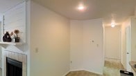 This single-floor space presents a clean, minimalist interior defined by smooth, white-painted walls and white baseboards that create a bright and open atmosphere. The image reveals a corner room bathed in warm light from a centrally placed recessed ceiling fixture, which highlights the textured ceiling finish overhead. The flooring features neutral-toned tile, providing a durable and easy-to-maintain surface that transitions toward a carpeted area visible in the adjoining hallway or room beyond. The walls are equipped with standard electrical outlets and a thermostat, indicating the space is climate-controlled for year-round comfort. A doorway or opening is visible to the right, suggesting a connected layout that allows for natural flow between areas. The overall design is straightforward and functional, offering a blank canvas that can be adapted to a variety of uses. The neutral color palette and simple finishes lend the space a clean, move-in-ready quality. The uncluttered, open feel of the room makes it well-suited for flexible use, whether as a living area, bedroom, or utility space.