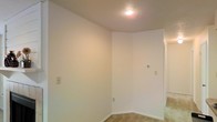 This single-floor space presents a clean, minimalist interior defined by smooth, white-painted walls and white baseboards that create a bright and open atmosphere. The image reveals a corner room bathed in warm light from a centrally placed recessed ceiling fixture, which highlights the textured ceiling finish overhead. The flooring features neutral-toned tile, providing a durable and easy-to-maintain surface that transitions toward a carpeted area visible in the adjoining hallway or room beyond. The walls are equipped with standard electrical outlets and a thermostat, indicating the space is climate-controlled for year-round comfort. A doorway or opening is visible to the right, suggesting a connected layout that allows for natural flow between areas. The overall design is straightforward and functional, offering a blank canvas that can be adapted to a variety of uses. The neutral color palette and simple finishes lend the space a clean, move-in-ready quality. The uncluttered, open feel of the room makes it well-suited for flexible use, whether as a living area, bedroom, or utility space.