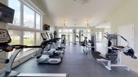 Located at 210 Highland Corporate Dr in Cumberland, Rhode Island, this fitness center offers a bright, well-appointed single-floor facility designed to support a comprehensive workout experience. The space is characterized by an abundance of natural light, courtesy of large, multi-pane windows lining the walls and offering views of the surrounding greenery and landscape.

The main workout area features durable rubber flooring and is equipped with a robust selection of cardio machines, including treadmills, ellipticals, and stationary bikes from reputable brands such as TRUE Fitness. Strength training is well-represented with a variety of cable and resistance machines, as well as free weights including dumbbells and a medicine ball. Wall-mounted flat-screen televisions provide entertainment during workouts.

Overhead, warm pendant light fixtures complement the natural light and contribute to the facility's clean, inviting atmosphere. The vaulted ceiling in sections of the space adds an open, airy quality throughout. A dedicated entry area with tile flooring and a reception-style counter provides a welcoming transition into the main exercise floor. The soft, neutral wall tones and crisp white trim enhance the overall bright and modern aesthetic, making this fitness center a functional and pleasant environment for exercise.