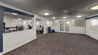 This fitness center is thoughtfully designed to accommodate a variety of exercise needs across two connected zones. The equipment area is outfitted with treadmills, a stationary bike, a weight bench, a dumbbell rack, and exercise balls, providing a comprehensive range of cardio and strength training options. White wainscoting panels line the lower walls, complemented by deep navy blue paint above, creating a clean and polished aesthetic. Patterned carpet in muted tones of brown and blue covers the floors throughout, adding warmth to the space. Ceiling fans with integrated lighting ensure comfortable air circulation, while flush-mount nautical-style light fixtures provide consistent illumination. Frosted glass double doors serve as a stylish entry point into the equipment area. The adjacent open studio space is generously proportioned, featuring a full-length wall mirror with a wooden ballet barre — ideal for stretching, fitness classes, or movement exercises. This area benefits from the same coordinated navy and white color scheme and offers ample open floor space for group activities. White structural columns divide the two areas subtly without interrupting the overall flow of the space.