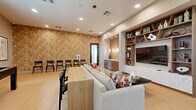 Located at 5900 N 99th Ave in Phoenix, AZ, this contemporary community lounge is thoughtfully designed to serve as a dynamic gathering and recreation space. The single-floor layout maximizes functionality through distinct activity zones, all unified by light wood-look flooring, recessed lighting, and a neutral color palette that creates a bright, welcoming atmosphere.

A standout feature is the bold geometric-patterned accent wall rendered in warm earthy tones, which anchors the space and adds significant visual character. Opposite, a recessed built-in shelving unit with a large flat-screen TV provides a media and entertainment focal point, complemented by decorative accessories and books.

The lounge area features a generous sectional sofa with colorful accent pillows, alongside a long communal wood table with black chairs — offering both relaxed seating and more structured collaborative use. A pool/game table positioned near large black-framed windows adds a recreational element, while natural light filters in through multiple windows with views of the surrounding building exterior.

A dedicated wet bar nook, finished in herringbone wood tile with pendant lighting, a countertop, and an under-counter refrigerator, adds practical entertaining amenities. Additional seating lines the accent wall, making the space well-suited for events or casual gatherings. Black-framed doors provide direct exterior access, further enhancing the open, connected feel of this well-appointed community amenity space.