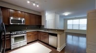 This single-floor residence showcases a well-designed open-concept layout where the kitchen flows seamlessly into the living area. The kitchen is a standout feature, equipped with rich medium-toned wood cabinetry that extends to the upper walls, providing generous storage. Granite countertops in warm, speckled tones run along the L-shaped counter configuration and extend to a peninsula with an integrated sink and dishwasher, creating a natural divide between the kitchen and living space. Stainless steel appliances, including a gas range with overhead microwave and a built-in dishwasher, lend a modern, functional aesthetic. A dark tile floor anchors the kitchen area, transitioning into warm hardwood flooring that extends throughout the open living space. Track lighting in the kitchen ensures ample task illumination, while a flush-mount fixture provides ambient light in the adjoining area. A large window in the living area invites natural light, brightening the overall space. Clean white walls and classic white trim complement the darker cabinetry and flooring, creating a balanced, inviting atmosphere throughout.