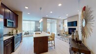 This thoughtfully designed single-floor residence offers a seamless open-concept layout that connects the kitchen, dining, and living areas into one expansive, light-filled space. Light hardwood flooring flows throughout, complementing the warm, neutral tones of the walls and creating a cohesive, inviting ambiance.

The kitchen is a standout feature, showcasing rich dark wood cabinetry, stainless steel appliances, and a generous center island topped with a white quartz countertop. Elegant pendant lighting hangs above the island, which offers bar seating — ideal for casual dining or entertaining. Abundant recessed lighting ensures the space is bright and functional at all times.

The living area is anchored by a fireplace and wall-mounted television, providing a comfortable focal point for relaxation. Large floor-to-ceiling windows with vertical blinds bathe the room in natural light while offering access to outdoor views. A secondary seating area further expands the generous living space.

The bedroom is a calm retreat, featuring recessed lighting, light hardwood floors, and direct access to a hallway. The bathroom is finished with dark wood vanity cabinetry, a white quartz countertop, a backlit mirror, and a full soaking tub with shower — combining modern style with everyday practicality throughout.