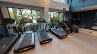 This modern fitness center is thoughtfully designed to support a comprehensive workout experience across a single, open-plan floor. The space is divided into distinct functional zones, each catering to different training styles. The cardio area is anchored by multiple treadmills and a StairMaster, positioned in front of large floor-to-ceiling windows that frame lush tropical greenery, flooding the space with natural light and creating an energizing atmosphere. Herringbone-patterned light wood flooring adds a refined aesthetic throughout this section, complemented by warm pendant lighting overhead.

The strength and free weights zone features adjustable workout benches, a power rack, and a well-stocked equipment wall with kettlebells, medicine balls, and a punching bag, set against a striking decorative panel. A wall-mounted television and a water fountain add practical amenities to this area.

A separate training alcove, finished in deep navy blue paneling, houses a rowing machine, an elliptical, and a boxing resistance machine with vibrant red pads. Full-length mirrors expand the sense of space visually. Recessed lighting throughout provides consistent, even illumination. The overall design balances a sleek, contemporary aesthetic with a functional, performance-oriented layout.
