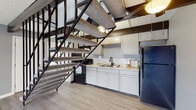 Located at 1503 D Monmouth Street in Lancaster, Ohio, this two-floor unit offers a distinctive blend of character and practicality. The main level features light luxury vinyl plank flooring throughout, complemented by dramatic exposed dark wood beam ceilings that add architectural depth to the living area. A bold, open-riser staircase with sleek black metal railings serves as a central design feature, connecting both floors while maintaining an open, airy feel. The kitchen is positioned conveniently beneath the staircase and is outfitted with gray cabinetry, granite-style countertops, a tile backsplash, double-basin sink, and black appliances including a range and refrigerator. A sliding glass door on the main level fills the space with natural light and provides outdoor access. The upper floor includes a carpeted bedroom with a ceiling fan, a built-in closet, and a window offering natural light. A full bathroom features a tub/shower combination with clean white surroundings. A dedicated laundry area accommodates a full-size washer and dryer alongside a water heater, adding everyday convenience. The overall palette of cool grays and crisp white trim creates a cohesive, modern aesthetic throughout the space.