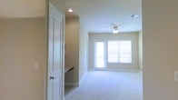 This single-story space is designed with a clean, functional layout that emphasizes openness and natural light. The main living area features soft, neutral-toned walls paired with plush wall-to-wall carpeting, creating a warm and inviting atmosphere. A ceiling fan with a built-in light fixture is centrally positioned, providing both airflow and ambient lighting to the room.

Natural light flows generously into the space through a large window fitted with white horizontal blinds, as well as a glass-paneled door that opens to an outdoor area, offering a seamless connection between the interior and exterior. The white trim work on the door and window frames adds a crisp contrast against the warm beige walls.

To one side of the space, a partial view reveals what appears to be a built-in countertop or workstation area, suggesting a dedicated functional zone separated by a classic white paneled door. Recessed lighting is visible in this adjoining area, adding layered illumination throughout. HVAC vents are integrated into the ceiling, ensuring climate control. Overall, the space presents a tidy, well-maintained environment with a straightforward, comfortable design aesthetic.