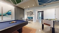 This single-floor game and entertainment lounge is thoughtfully designed to encourage social interaction and recreation. The space is defined by its striking architectural ceiling treatments, which feature geometric coffered panels accented in bold colors—green, blue, and orange—adding a dynamic visual element overhead. Recessed lighting throughout ensures the room is well-illuminated.

The main gaming area houses a full-size pool table with a blue felt surface, a Tornado foosball table, and a dedicated poker room featuring a round gaming table surrounded by patterned upholstered chairs. A popcorn machine stationed nearby adds to the leisure atmosphere. Large windows in the poker area allow natural light to filter in and provide views to the exterior.

A built-in shelving and media wall finished in deep navy blue anchors one side of the room, offering display storage and a mounted television screen. A fluted vertical accent wall provides textural contrast alongside large abstract artwork panels. A cozy lounge nook with a bold bullseye graphic mural, teal banquette seating, and a small café table creates an intimate conversational corner. Polished concrete flooring with area rugs ties the various activity zones together seamlessly.