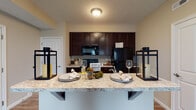 Located at 7220 E Broad St in Columbus, Ohio, this single-floor residence offers a thoughtfully designed layout with a seamless blend of functionality and comfort. The open-concept kitchen is a standout feature, equipped with rich dark-espresso cabinetry reaching toward the ceiling, light granite-style countertops, and black appliances including a range, over-the-range microwave, and refrigerator. A breakfast bar peninsula provides additional seating and counter space, making it ideal for casual dining or entertaining. The adjacent living area is generously sized, featuring durable wood-style flooring that transitions into plush carpet, and a sliding glass door fitted with vertical blinds that invites natural light while providing access to the outdoors. The bedroom is carpeted for warmth and comfort and includes a closet with built-in shelving, offering ample storage. The full bathroom is well-appointed with a tub and shower combination, a dark-cabinetry vanity with a granite-style countertop, a large mirror, and tile flooring. Neutral warm tones throughout the residence create a cohesive and welcoming ambiance, making this a practical and comfortable home in a convenient Columbus location.