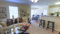 Located at 1525 E Pass Rd in Gulfport, Mississippi, this single-story home offers a comfortable and well-appointed layout designed for easy living. The open-concept floor plan seamlessly connects the living room and dining area, creating a spacious and cohesive environment ideal for both everyday living and entertaining. The living room features plush carpeting, crown molding, and a ceiling fan, while large windows fitted with white plantation-style blinds allow generous natural light to fill the space. A glass-top coffee table and coordinating furnishings add a contemporary touch to the warm, neutral color palette of the walls and flooring. The adjacent dining area is highlighted by a classic chandelier pendant light and a dark wood dining set, continuing the open and airy feel. A pass-through counter connects the dining space to the kitchen, enhancing the home's functional flow. Throughout the interior, thoughtful decorative details — including candle accents, artwork, and pops of color — contribute to an inviting and well-styled ambiance. The seamless transition between living spaces and the abundance of natural light make this Gulfport home a bright and welcoming retreat.
