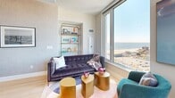 Perched atop 1515 Surf Ave in Coney Island, Brooklyn, Penthouse 3C is a three-bedroom, one-bathroom residence offering breathtaking panoramic views of the Atlantic Ocean, Coney Island beach, and the Manhattan skyline. Light-toned hardwood floors run throughout, complementing the bright, white interior walls and crisp modern aesthetic.

The living area is anchored by dramatic floor-to-ceiling corner windows that flood the space with natural light and frame sweeping waterfront views. A streamlined kitchen with warm wood cabinetry and stainless steel appliances sits adjacent to the living space. A cozy secondary lounge area features a built-in shelving niche and its own large window overlooking the beach below.

Each of the three bedrooms offers its own distinct character. The primary bedroom is generously proportioned with a bold floral accent wall, large windows with open city views, and mirrored nightstands. A second bedroom features sliding closet doors and elevated ceilings, while the third bedroom benefits from a bright corner window with urban vistas and built-in wall sconces. All bedrooms include closet storage with shelving systems.

The bathroom is finished in large-format marble-look tiles, a soaking tub with shower, a floating wood-toned vanity, and an illuminated mirror, delivering a clean, spa-like atmosphere.