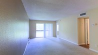 This single-floor space presents a straightforward and functional layout centered around a generously sized living area. The room is finished with wall-to-wall carpeting in a neutral tone, providing a soft underfoot feel that extends throughout the open floor plan. Walls are painted in a warm beige, contributing to a bright and understated ambiance.

A prominent sliding glass door fitted with vertical blinds serves as the focal point of the main living area, allowing ample natural light to fill the room while providing access to an outdoor balcony. The view through the glass hints at surrounding greenery, adding a subtle connection to the outdoors.

The textured popcorn ceiling adds a classic residential character to the space, while white baseboards frame the perimeter of the room cleanly. A doorway visible toward the rear of the living area suggests additional rooms beyond, indicating a connected layout designed for ease of movement.

Overall, the space maintains a clean, neutral palette that offers flexibility for a variety of furnishing arrangements, making it a practical and adaptable environment for everyday living.