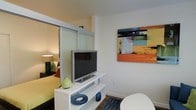 This single-floor space showcases a thoughtfully designed open-concept layout that maximizes functionality within a compact footprint. A key architectural feature is the frosted glass sliding partition, which elegantly divides the living area from the sleeping zone while maintaining a sense of openness and allowing light to filter between the two spaces.

The living area is anchored by a sleek white media console, which houses a flat-screen television and provides additional display and storage space. Decorative accents, including ceramic pieces in navy and green tones, add subtle pops of color to the clean, white surfaces. A colorful abstract canvas artwork adorns the wall, introducing vibrant shades of orange, blue, green, and purple that energize the space.

Dark hardwood-style flooring runs throughout, providing a warm contrast to the predominantly white walls and furnishings. The sleeping area, visible through the translucent partition, features warm ambient lighting from a bedside lamp, contributing to a cozy, intimate atmosphere.

Overall, the space presents a modern, urban aesthetic with clean lines, a neutral base palette accented by bold decorative touches, and smart design solutions that balance privacy with an open feel.