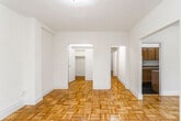 This single-floor residence is characterized by warm parquet wood flooring laid in a distinctive basketweave pattern that runs consistently throughout every room, lending a cohesive and classic aesthetic to the entire space. The open layout connects a spacious living area to the rest of the unit through a combination of arched and rectangular doorways, creating an inviting flow between rooms.

The living area is generously proportioned, featuring neutral cream-toned walls, clean white baseboards, and a radiator unit that provides efficient heating. Corner windows in one of the bedrooms allow natural light to enter from multiple angles, offering views of surrounding greenery and the exterior streetscape. Each bedroom is well-sized, with one featuring a dedicated walk-in closet equipped with built-in shelving rods, and another providing direct access to the bathroom.

The kitchen is outfitted with rich, dark-stained wood cabinetry offering ample upper and lower storage, stone-finish laminate countertops, a built-in sink, and a full-size refrigerator. Dark speckled tile flooring grounds the kitchen's design. The bathroom features classic white subway tile wainscoting, mosaic tile flooring, a soaking tub with shower, and a window for natural ventilation.