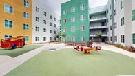 Located at 3515 Vista Ln in San Diego, California, this outdoor courtyard is a dynamic communal space defined by its striking architectural surroundings and thoughtfully designed amenities. The multi-story buildings enclosing the courtyard feature bold, eye-catching facades in a vivid palette of red, orange, magenta, teal, green, yellow, and white — creating a visually energetic environment that stands out as a defining characteristic of the property.

The courtyard is organized into distinct functional zones. A children's play area features soft rubber surfacing in a flowing green layout, equipped with colorful play structures including a fire truck climber and spring-mounted ride-on toys, providing a safe and engaging space for young residents. Adjacent to the play area, wide concrete pathways connect multiple seating zones furnished with durable metal benches, offering residents comfortable spaces to gather and socialize.

Landscaping throughout the courtyard includes young trees, native ornamental grasses, and shrub plantings set in mulched beds along the building perimeters, adding natural texture and greenery to the hardscape. The open-air design allows for ample natural light, with the surrounding multi-story buildings framing the sky above. Covered walkways and balconies visible on the upper floors further reinforce the sense of an active, well-connected residential community within this San Diego location.