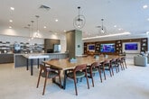 This single-floor amenity space in Charleston, SC, showcases a cohesive modern design highlighted by light-toned large-format tile flooring throughout. The welcoming lounge entry features a bold, colorful abstract artwork as a focal point, complemented by a mix of cognac leather and neutral upholstered seating arranged around accent tables. A decorative damask-patterned wallcovering adds texture to one wall, creating visual interest.

The kitchen area is well-appointed with a substantial island featuring paneled blue-gray cabinetry and quartz countertops, stainless steel appliances, and a striking elongated hexagonal tile backsplash. A dark lattice-style screen partition adds a layer of visual depth, while a separate galley-style kitchenette with a coffee station serves additional needs.

The media lounge offers generous seating around a large wall-mounted screen framed by rich wood built-in shelving with display niches. Cove lighting along the tray ceiling and a vibrant patterned area rug contribute warmth and comfort.

A conference or dining area features a substantial solid wood table surrounded by chairs, with floor-to-ceiling black-framed glass doors and windows providing abundant natural light and direct access to an outdoor terrace area.