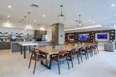 This single-floor amenity space in Charleston, SC, showcases a cohesive modern design highlighted by light-toned large-format tile flooring throughout. The welcoming lounge entry features a bold, colorful abstract artwork as a focal point, complemented by a mix of cognac leather and neutral upholstered seating arranged around accent tables. A decorative damask-patterned wallcovering adds texture to one wall, creating visual interest.

The kitchen area is well-appointed with a substantial island featuring paneled blue-gray cabinetry and quartz countertops, stainless steel appliances, and a striking elongated hexagonal tile backsplash. A dark lattice-style screen partition adds a layer of visual depth, while a separate galley-style kitchenette with a coffee station serves additional needs.

The media lounge offers generous seating around a large wall-mounted screen framed by rich wood built-in shelving with display niches. Cove lighting along the tray ceiling and a vibrant patterned area rug contribute warmth and comfort.

A conference or dining area features a substantial solid wood table surrounded by chairs, with floor-to-ceiling black-framed glass doors and windows providing abundant natural light and direct access to an outdoor terrace area.
