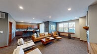 This well-appointed single-floor community space offers a thoughtfully designed interior that blends mid-century modern aesthetics with functional amenities. The main lounge area serves as the centerpiece, featuring rich cognac leather sofas and accent chairs arranged around a solid wood coffee table, all anchored by plush carpeting. A gas fireplace set into a teal accent wall provides a warm focal point, complemented by a large wall-mounted flat-screen television and a coordinating wood media console. A shuffleboard table positioned near the windows adds a recreational element to the space.

Natural light flows generously throughout via multiple large windows fitted with blinds, offering views of the surrounding greenery. The fully equipped kitchen boasts warm wood cabinetry, a mosaic tile backsplash, white countertops, stainless steel appliances including a range and microwave, and a curved island with bar seating — ideal for casual entertaining. Dark hardwood flooring grounds the kitchen area, transitioning to carpet throughout the lounge.

A dedicated dining nook provides additional seating with round glass-top tables and upholstered chairs, framed by curated wall art and elegant dried floral arrangements. The overall ambiance is modern, inviting, and well-suited for both social gatherings and relaxed everyday use.
