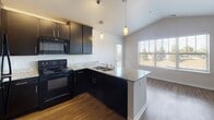 Located at 3111 Market Ln in Kenosha, Wisconsin, this single-floor unit offers a well-organized layout with a clean, modern aesthetic throughout. The kitchen serves as a central highlight, featuring dark espresso-toned cabinetry, granite countertops, a peninsula with a double sink, pendant lighting, and black Whirlpool appliances including a range and over-the-range microwave. Natural light filters in through a large window adjacent to the kitchen, brightening the connected living and dining area. The living space is finished with warm hardwood-style plank flooring and a ceiling fan with integrated lighting, creating a comfortable and functional environment. A half-wall partition with a open pass-through connects the lower living area to the upper level, adding an architectural detail that enhances the sense of openness between spaces. The bathroom is equipped with a single-sink vanity with dark cabinetry, a large mirror, and a full tub-shower combination, finished with tile-style flooring. Neutral wall tones and white trim are consistent throughout, contributing to a cohesive and understated interior palette. The overall design prioritizes practicality while maintaining a modern, move-in-ready appeal.