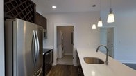 This single-floor space showcases a thoughtfully designed kitchen that serves as its centerpiece. The kitchen features dark espresso-toned cabinetry, including a distinctive wine rack unit that adds both functionality and character. Stainless steel appliances, including a French door refrigerator, microwave, and range, provide a sleek, contemporary aesthetic that contrasts elegantly with the dark cabinetry. A generous island or peninsula with a crisp white quartz countertop offers ample workspace, complemented by an undermount stainless steel sink and a modern gooseneck faucet. Pendant lighting fixtures hang above, adding warmth to the space alongside recessed ceiling lights. The rich, dark hardwood flooring runs throughout, grounding the design with a sense of warmth and continuity. A dedicated laundry room is conveniently accessible directly from the kitchen, housing a stacked washer and dryer unit — a practical and space-efficient feature. White trim and neutral wall tones throughout create a clean, balanced backdrop that enhances the overall brightness and modern sensibility of the space.