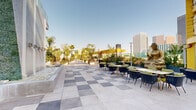 This expansive rooftop terrace offers a sophisticated outdoor retreat set high above a bustling urban downtown. The space is defined by a distinctive checkered tile pattern spanning the open-air floor, creating a bold visual foundation throughout. A dramatic centerpiece living green wall, adorned with cascading water features, anchors one end of the terrace and serves as a striking architectural focal point.

The terrace is thoughtfully divided into several zones. A dining and lounge area features contemporary dark chairs with green cushions arranged around sleek tables, ideal for gatherings against the backdrop of a gleaming city skyline. A lush lawn area bordered by vibrant flowering plants, palm trees, and colorful rose hedges provides a softer, garden-like retreat. String lights overhead lend a warm, ambient character to this zone.

A more open, flexible section accommodates recreation and events, featuring lawn games such as cornhole and an oversized chess set. Perimeter walkways lined with blooming flower planters offer scenic promenades with unobstructed skyline views. Glass railings along elevated sections ensure panoramic sightlines without visual interruption. The overall ambiance balances urban energy with natural serenity, making this terrace a dynamic and versatile outdoor destination.