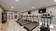 Located at 525 Stone Pointe Dr in Manhattan, Kansas, this fitness center is a thoughtfully designed facility spread across a single floor. The space is organized into three distinct exercise rooms and a connecting hallway, providing a structured and functional layout for a variety of workout routines.

The primary exercise area houses a row of True-brand treadmills and an elliptical trainer, all positioned on protective floor mats atop light-toned, wood-look luxury vinyl flooring. Wall-mounted flat-screen televisions are positioned for easy viewing during cardio sessions. Full-length framed mirrors and a freestanding shelving unit add both practicality and depth to the room. Recessed ceiling lighting throughout provides consistent, well-distributed illumination.

A second exercise room is dedicated to strength training, featuring a multi-station cable machine that offers a range of resistance exercises. This room benefits from generous open floor space, allowing for free-movement workouts alongside the equipment.

The third exercise room provides a quieter, more focused environment, featuring a recumbent exercise bike and a window that introduces natural light. Neutral warm-gray walls and clean white baseboards run consistently throughout all rooms, creating a cohesive and modern aesthetic. The overall design is clean, practical, and welcoming, well-suited for daily fitness use.