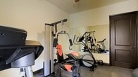This single-floor fitness space is thoughtfully outfitted to support a well-rounded exercise routine. The room features a multi-function home gym station, ideal for strength training across a variety of muscle groups, alongside a cardio machine for aerobic workouts. Kettlebells are available for free-weight training, adding further versatility to the space. A large wall-mounted mirror spans a significant portion of the room, enhancing the sense of openness while allowing users to monitor their form during exercise. The neutral-toned walls and concrete-style flooring give the space a clean, functional aesthetic typical of a dedicated workout environment. A ceiling fan provides ventilation and airflow, keeping the area comfortable during intense training sessions. The layout is efficiently organized to maximize usable floor space while keeping equipment accessible. Whether used for strength training, cardio, or flexibility work, this fitness space provides a practical and focused environment for daily exercise.